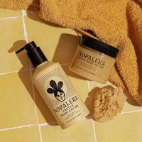 Nopalera Cactus Body Cream and Cactus Body Polish in a warm yellow tiled setting with a folded mustard towel and a natural loofah sponge.