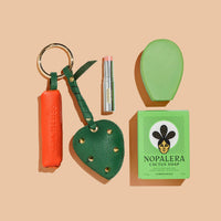 Flat lay of Nopalera products on a beige background, featuring a cactus soap bar with packaging, a green soap dish, a lip balm, and two leather accessories with key rings.
