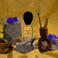 Perfume bottle labeled 'Dulce de Cuerpo' with incense sticks, flowers, and vanilla beans on a yellow background.