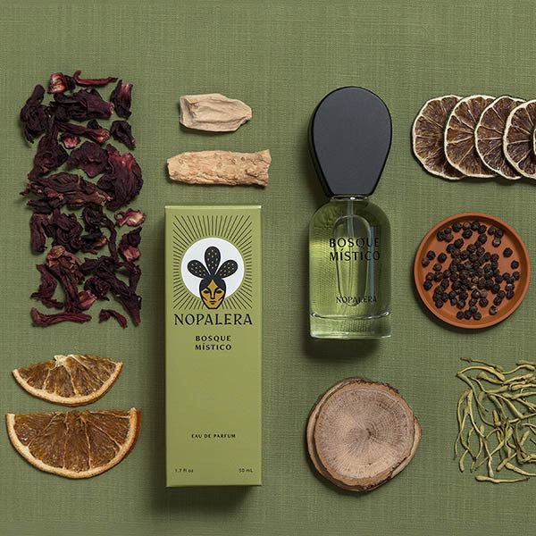 A styled flat-lay of a Nopalera Bosque Místico Eau de Parfum bottle and its green box, surrounded by dried botanical ingredients including hibiscus, citrus slices, cinnamon, and peppercorns on an olive green textured background.