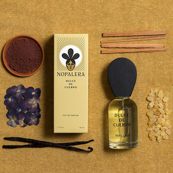 Nopalera Dulce de Cuerpo eau de parfum bottle and its packaging box, styled with natural ingredients (vanilla beans, cinnamon sticks, purple flowers, and dried roots) on a textured tan linen background.