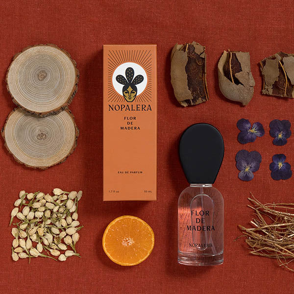 Nopalera Flor de Madera Eau de Parfum bottle and box arranged on a rust-red fabric background with botanical props including wood slices, dried bark, jasmine blossoms, a sliced orange, and purple flowers.