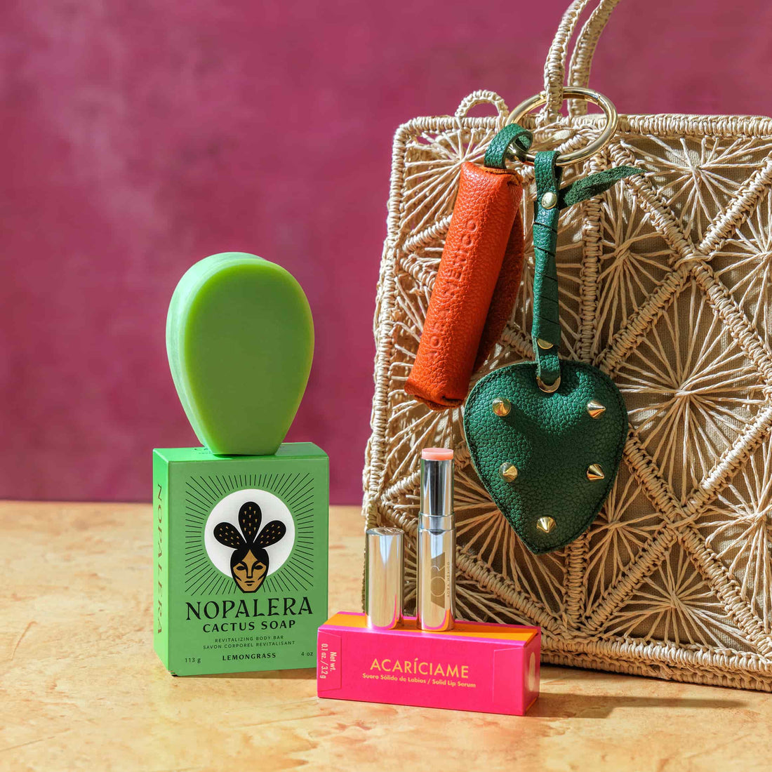 Nopalera cactus soap with packaging, green soap dish, lip balm, and leather keychain accessories displayed next to a woven handbag against a pink background.