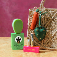 Nopalera cactus soap with packaging, green soap dish, lip balm, and leather keychain accessories displayed next to a woven handbag against a pink background.