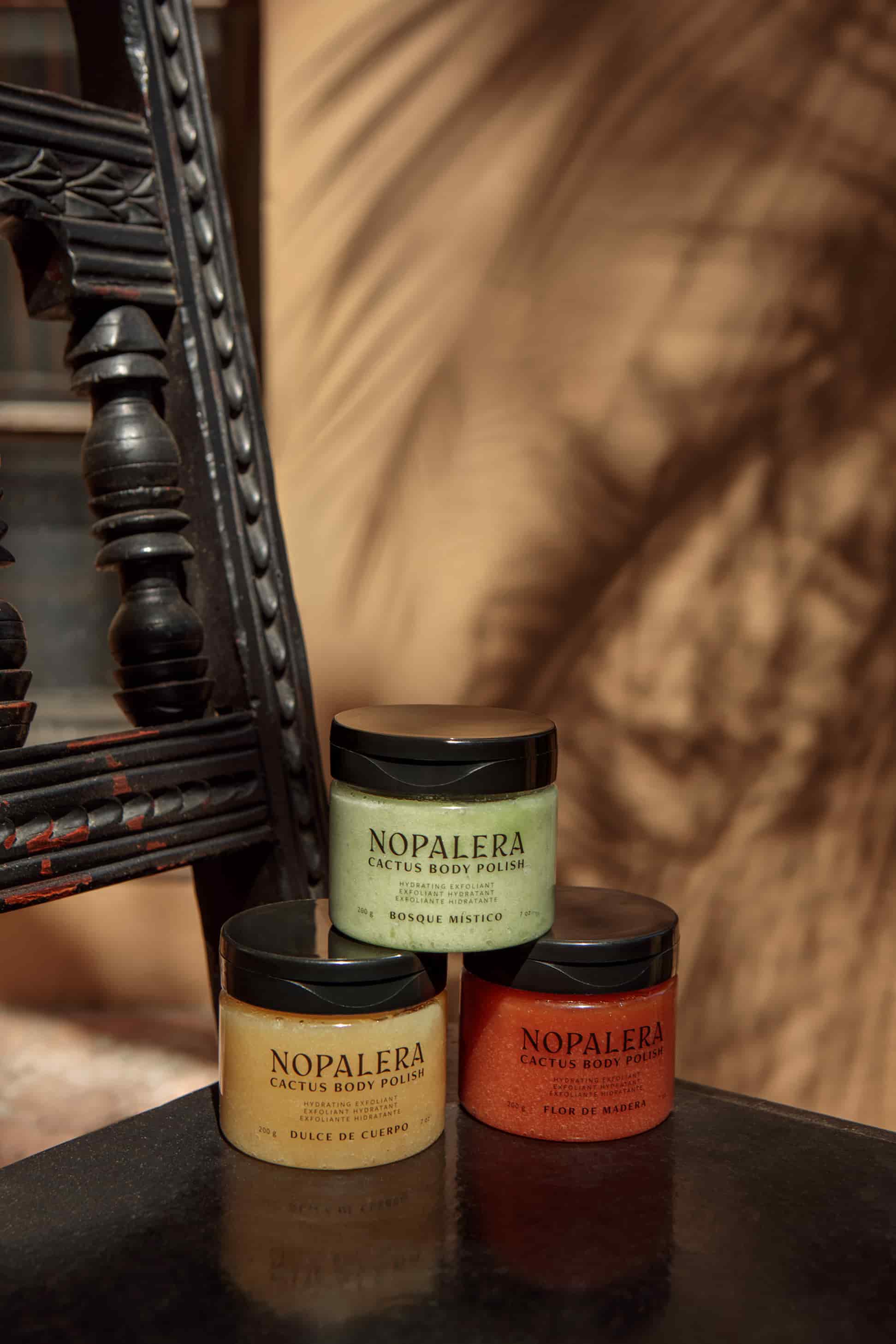 Three Nopalera Cactus Body Polish jars arranged on a dark reflective surface next to an ornate black wooden chair, with a warm brown draped fabric background.