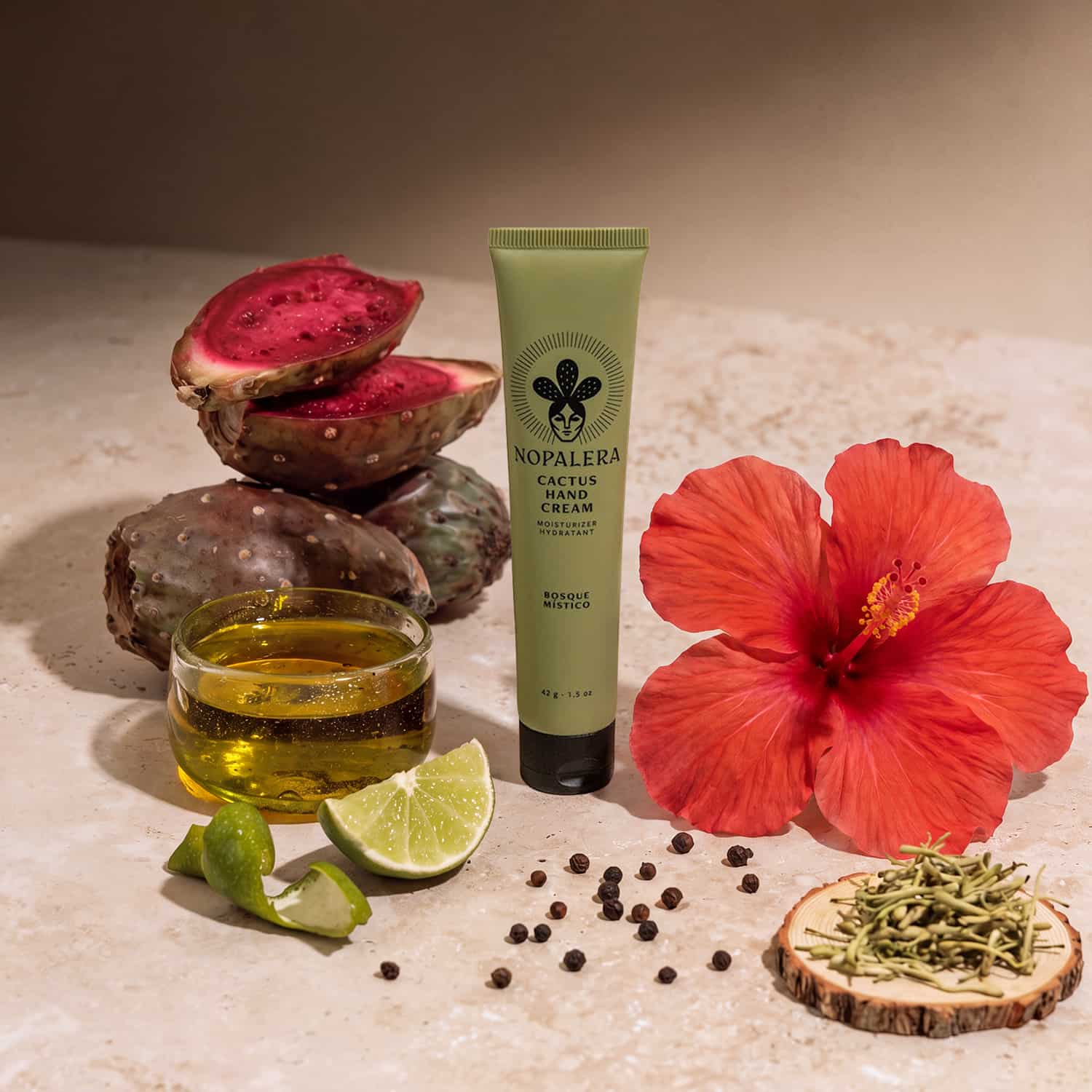 A tube of Nopalera Cactus Hand Cream stands on a stone surface surrounded by natural ingredients: sliced red cactus, green limes, a small glass of yellow oil, black peppercorns, dried herbs on a wood slice, and a large red hibiscus flower.