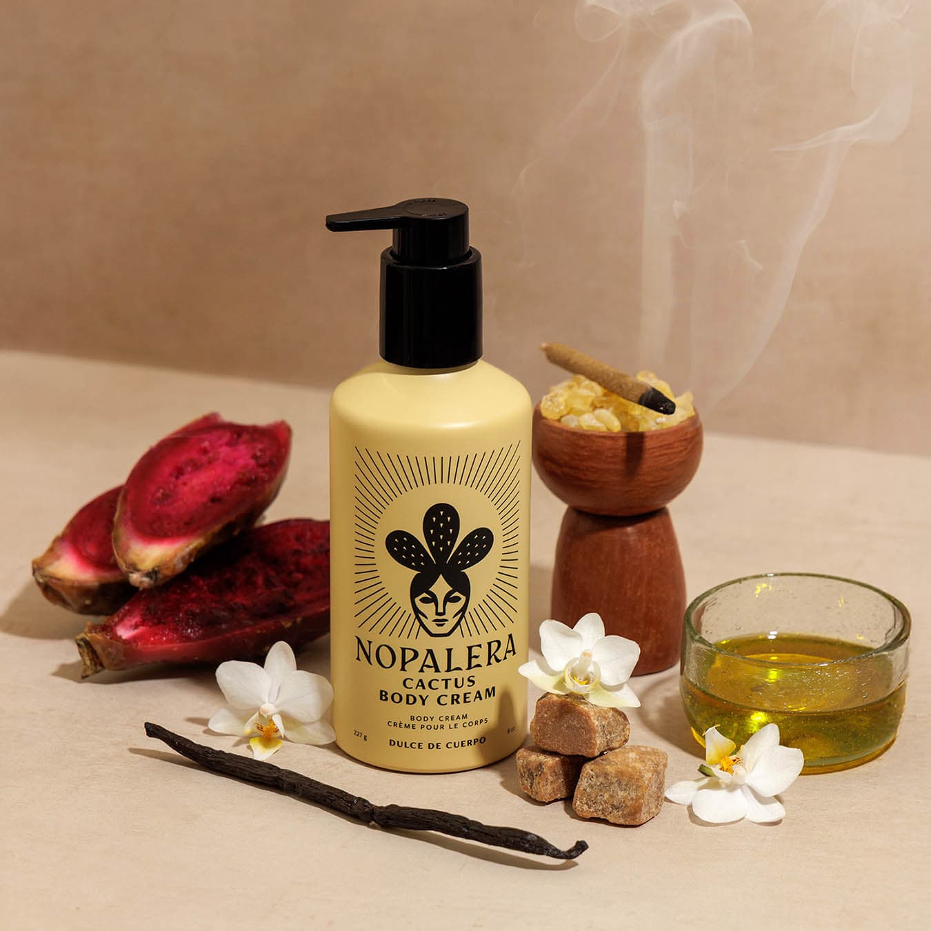 A bottle of Nopalera Cactus Body Cream stands on a beige surface with styled natural ingredients around it, including sliced red cactus, white flowers, brown sugar cubes, a vanilla bean, a small bowl of yellow liquid, and a wooden grinding tool with smoke rising in the background.