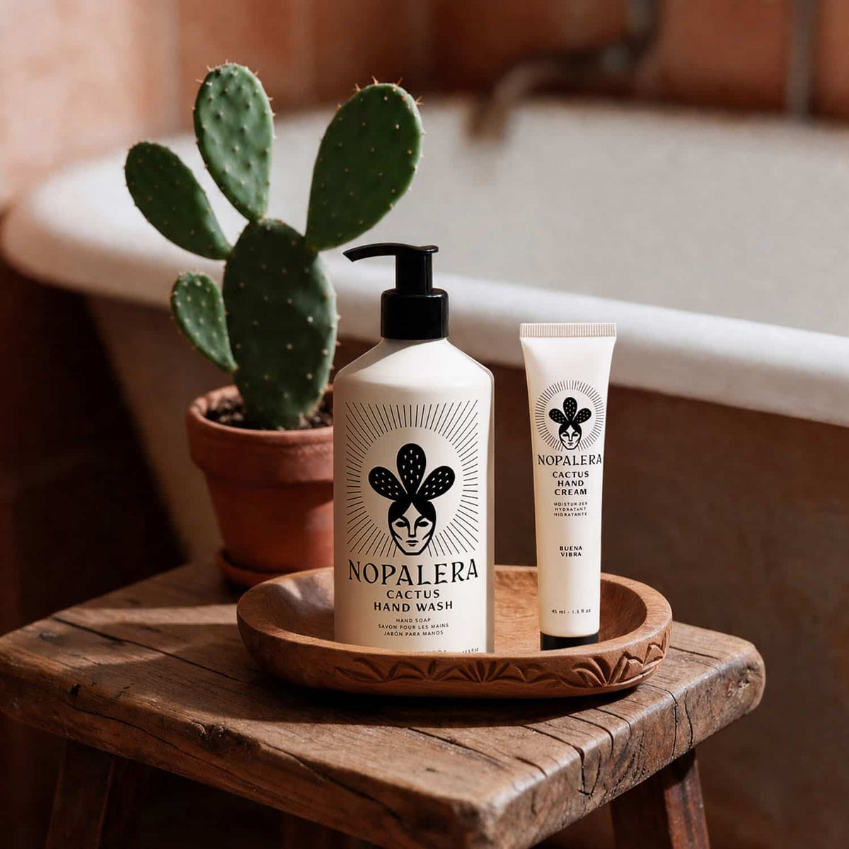 Nopalera Cactus Hand Wash and Cactus Hand Cream in Buena Vibra scent on a carved wooden tray, set on a rustic stool beside a potted prickly pear cactus and a bathtub.