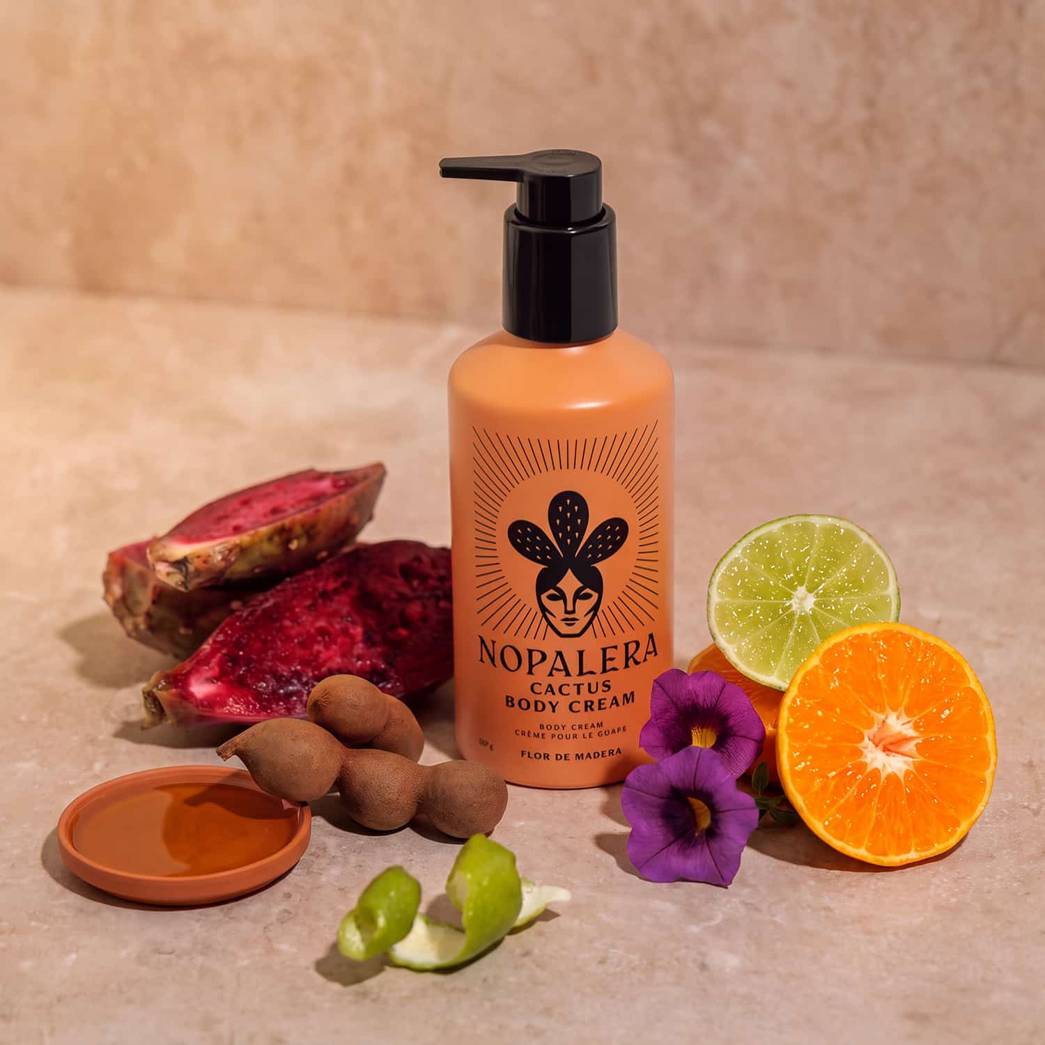 A bottle of Nopalera Cactus Body Cream stands on a beige stone surface surrounded by natural ingredients including a sliced orange, lime, purple flowers, a small dish of honey, and dried fruit pieces. The bottle is matte orange with a black pump and a black printed logo.