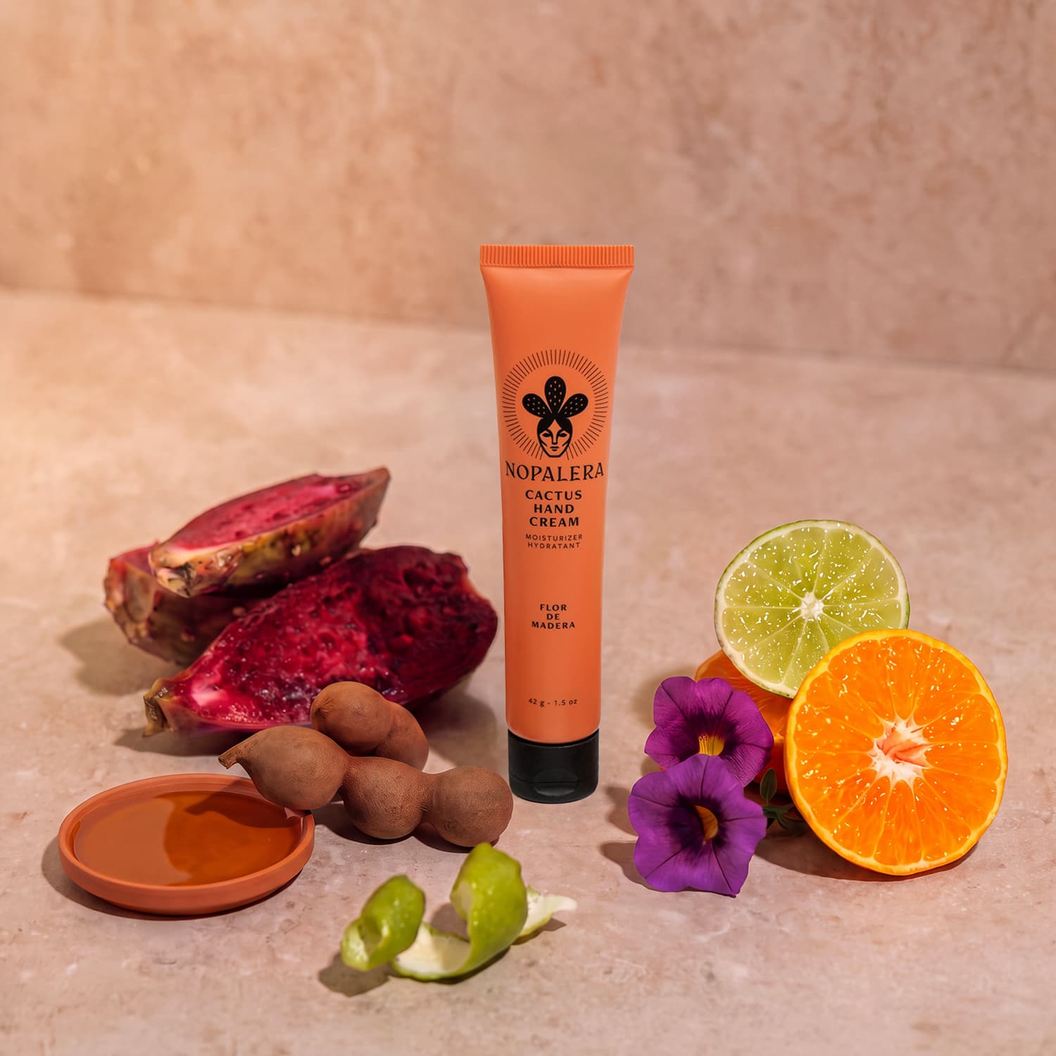 An orange tube of Nopalera Cactus Hand Cream stands upright on a beige stone surface, surrounded by natural ingredients including a sliced red dragon fruit, lime halves, an orange half, purple flowers, a small dish of honey, and a green lime peel.