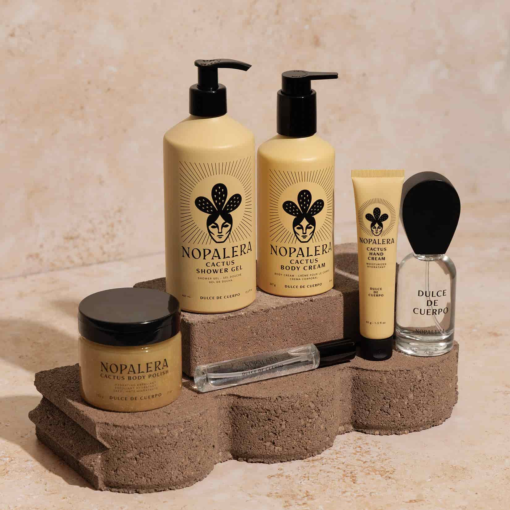 A set of Nopalera Dulce de Cuerpo skincare and fragrance products arranged on rustic stone blocks against a textured beige wall. The products include a cactus body polish jar, two pump bottles of shower gel and body cream, a hand cream tube, a clear perfume bottle labeled Dulce de Cuerpo, and a travel spray.