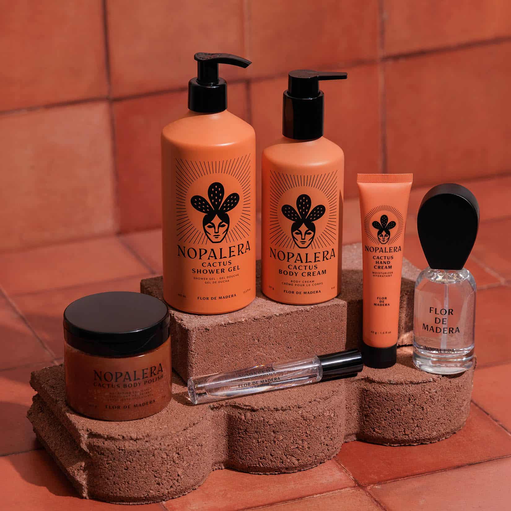 A coordinated set of Nopalera flor de Madera skincare and fragrance products arranged on stacked terracotta blocks against a terracotta tile background. Visible items include two large pump bottles labeled Cactus Shower Gel and Body Cream, a smaller tube labeled Hand Cream, a clear glass bottle labeled Flor de Madera  perfume a brown jar labeled body polish, and a clear travel spray perfume with a black cap.