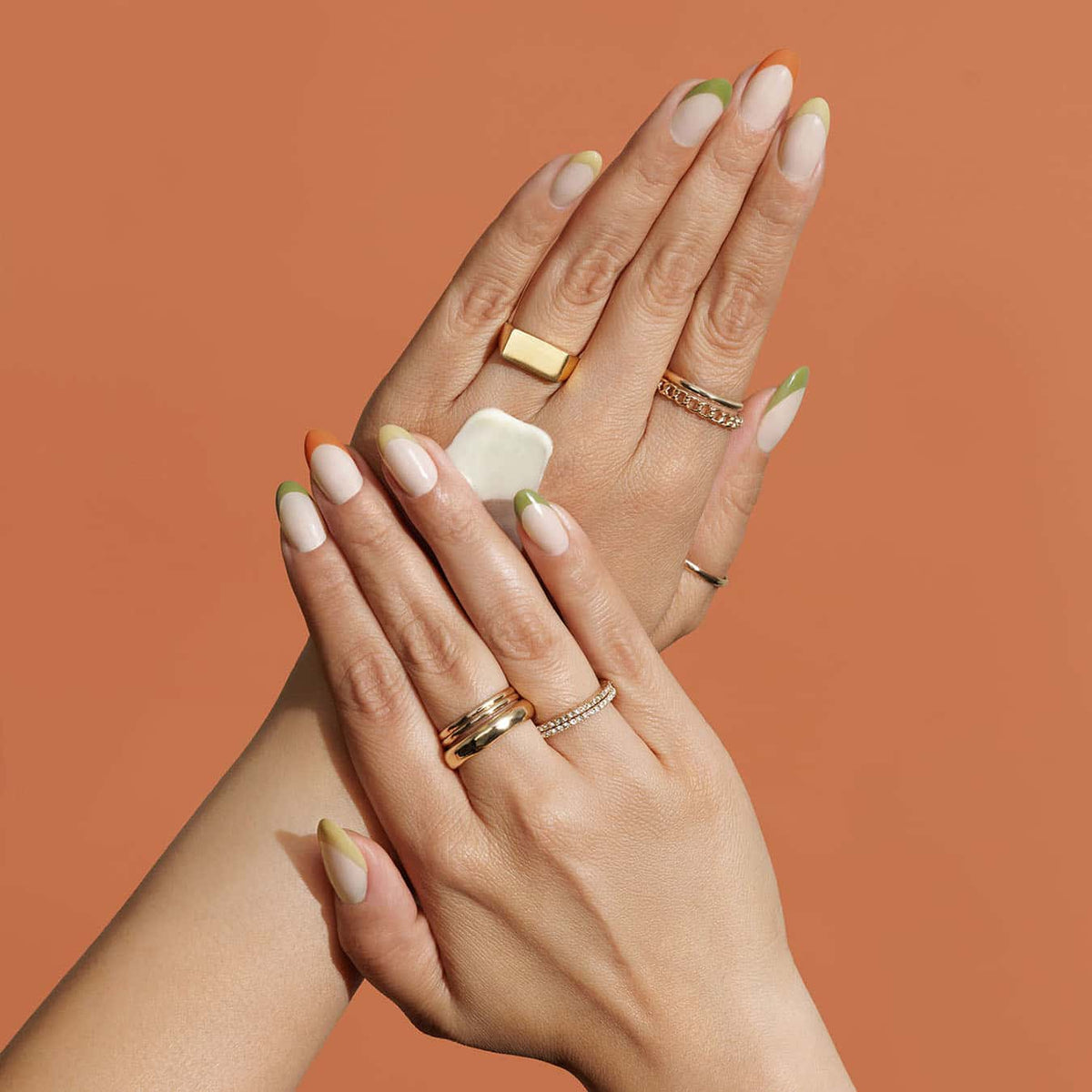 Two hands with manicured nails posed against a solid terracotta background, with one hand gently applying and massaging cream into the other. The hands are adorned with multiple gold rings, including a large square-cut stone ring, a diamond-accented band, and a thin chain ring.