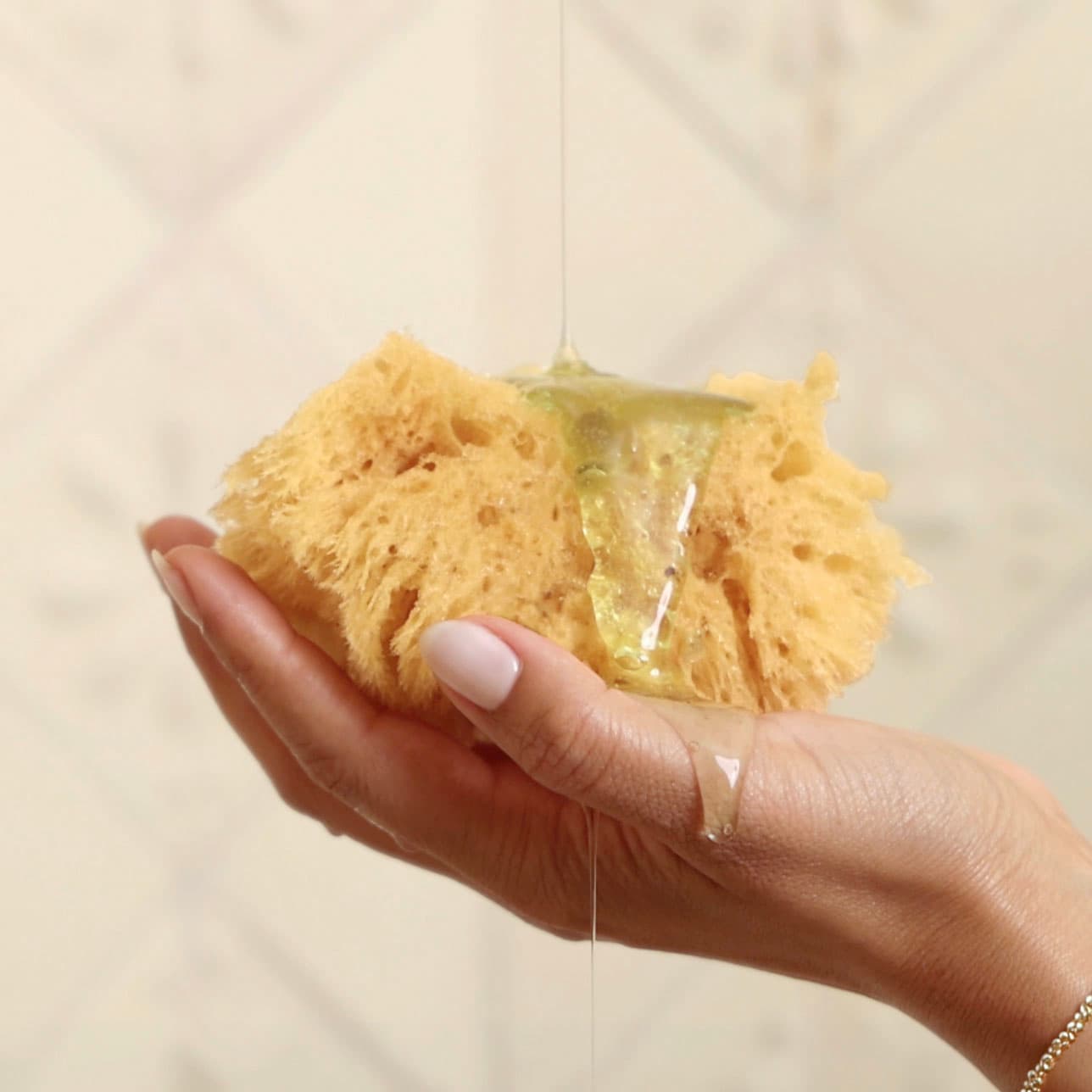 A hand holding a yellow porous sponge with a clear gel or liquid dripping from it, against a softly blurred beige tiled background.