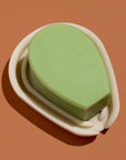 A green cactus-shaped bar of soap resting in a white silicone soap dish against a brown background.