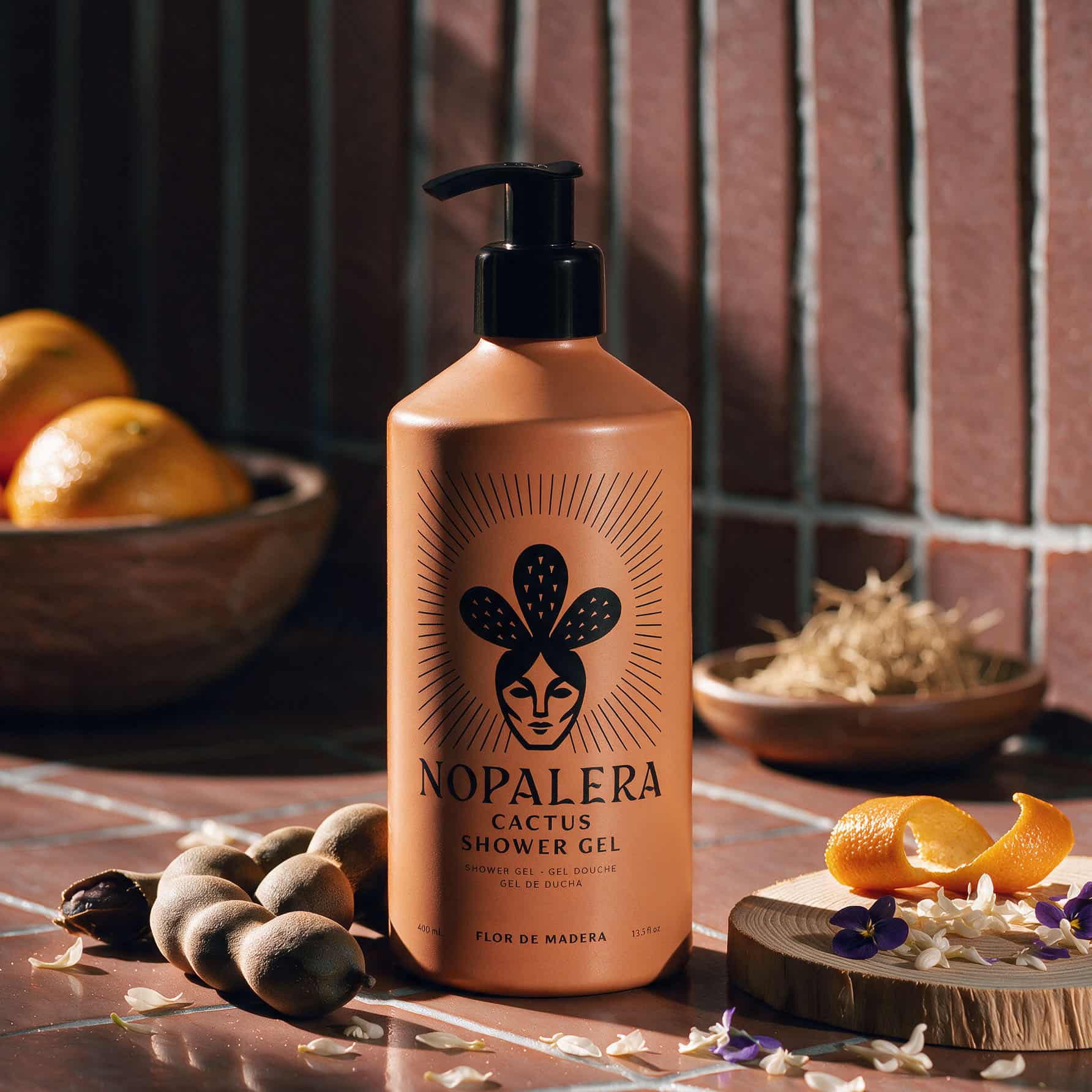 A bottle of Nopalera Cactus Shower Gel stands on a tiled surface with dried cactus pieces, citrus peels, and small flowers around it. The bottle is terracotta-colored with a black pump and black printed logo, set against a rustic background with a bowl of oranges and a brick wall.