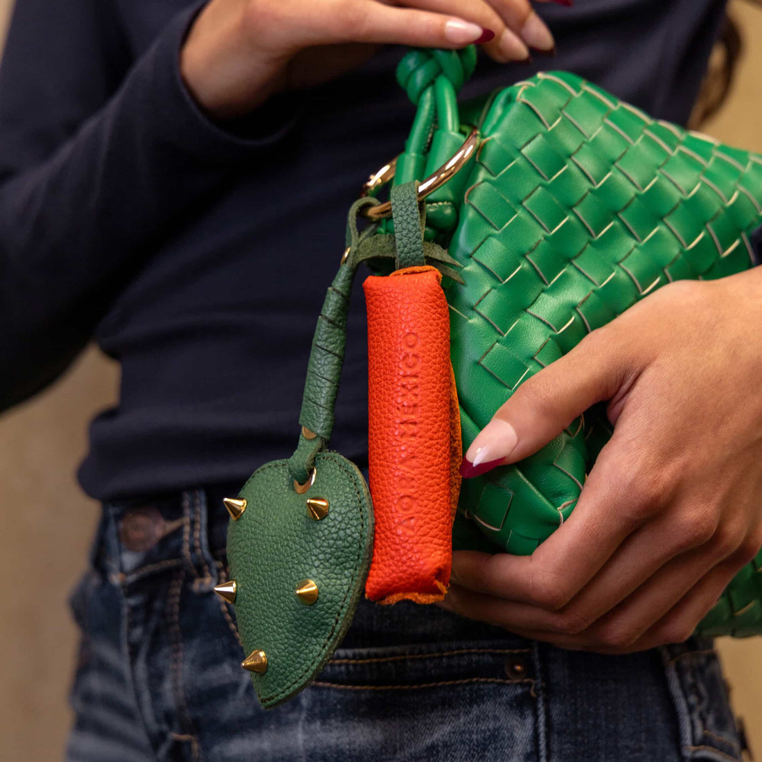 Close-up of a person holding a green woven handbag with Nopalera leather keychain accessories attached, including a green cactus-shaped charm with gold studs and an orange leather charm.