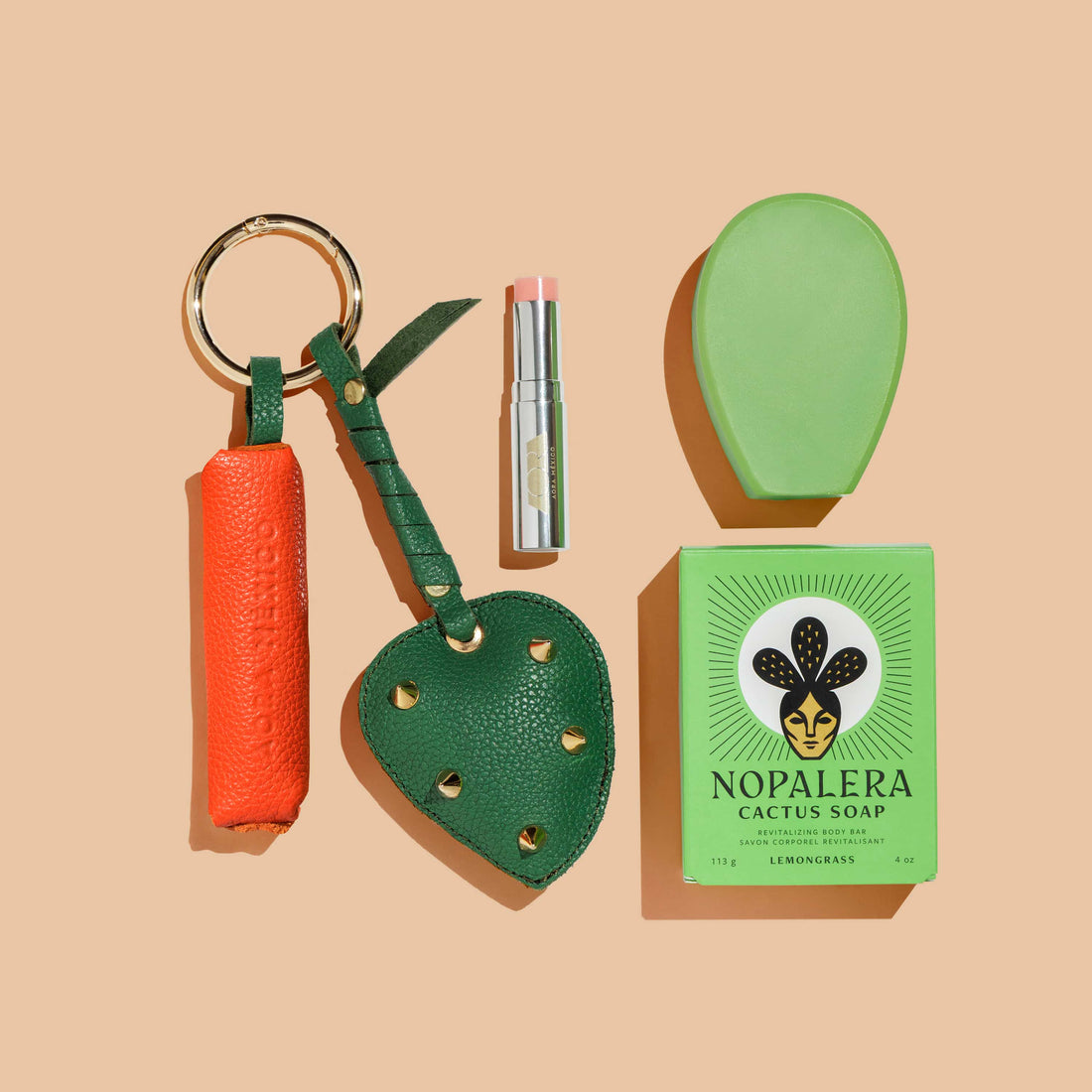 Flat lay of Nopalera products on a beige background, featuring a cactus soap bar with packaging, a green soap dish, a lip balm, and two leather accessories with key rings.