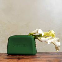Green Nopalera cosmetic pouch with gold zipper and embossed logo on a neutral background and flowers