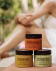 Three jars of Nopalera body polish stacked on a surface with a blurred person sitting outdoors in the background.