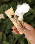 Hand holding a tube of Nopalera cactus hand cream Dulce de Cuerpo with a white flower in the background.