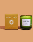 A lit candle with a green container, labeled 'Nopalera Todos Santos', next to its packaging box with the same brand name and floral design.
