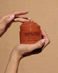 Hand holding a jar of Nopalera cactus body polish Flor de Madera against a beige background