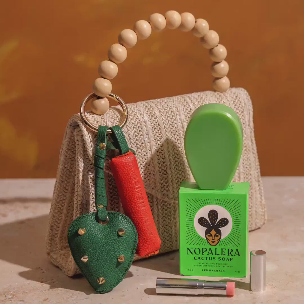 Short product video showing a woven handbag with beaded handle and Nopalera cactus soap, a close-up of hands attaching colorful leather keychain accessories to a bag, and a woman applying lip balm for a natural beauty routine.